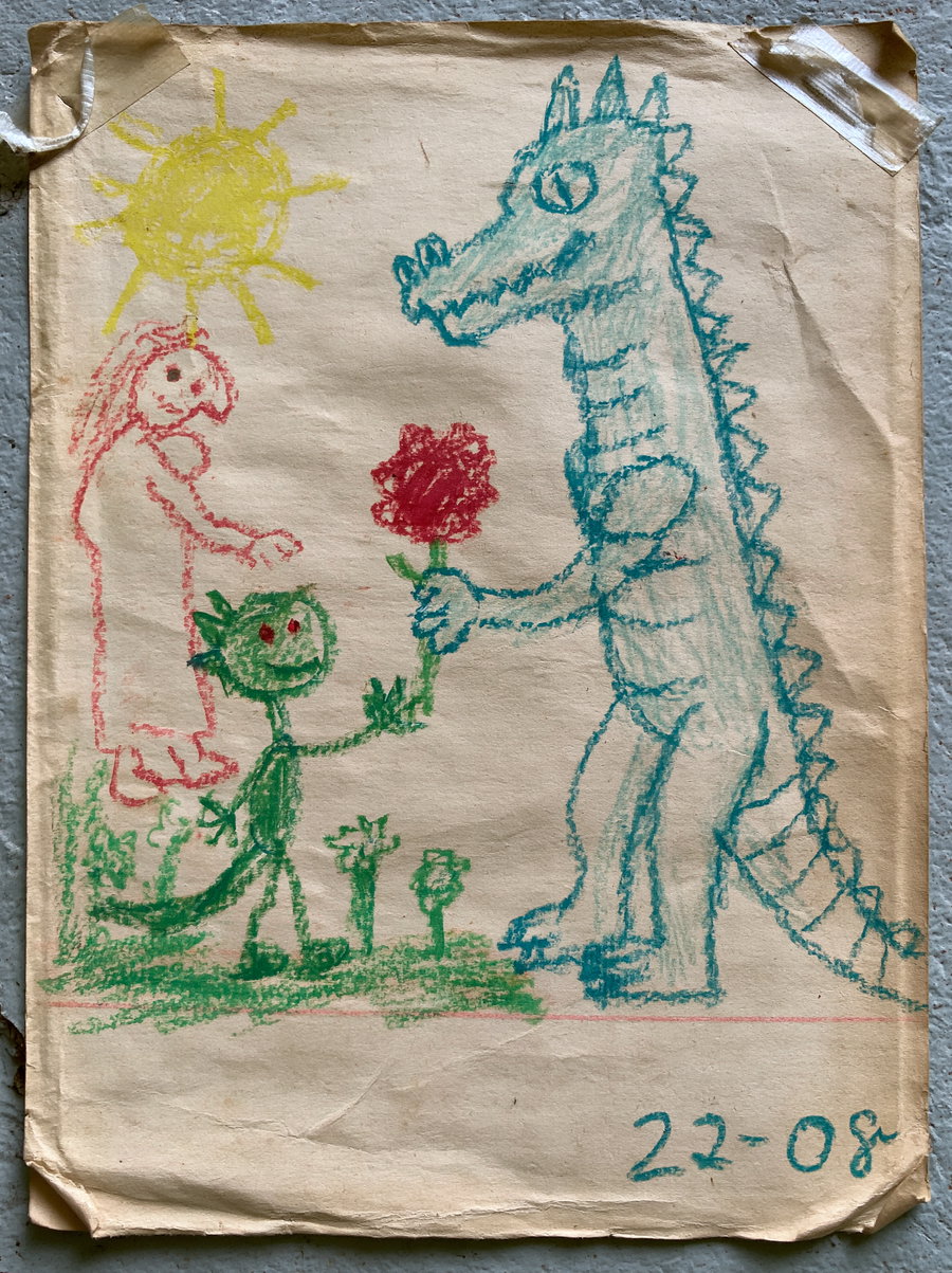 Crayon drawing on paper showing a green dragon holding out a red flower to a larger blue dragon standing on two legs, with a person figure and a sun drawn in yellow in the top left corner, plus some grassy plants at their feet