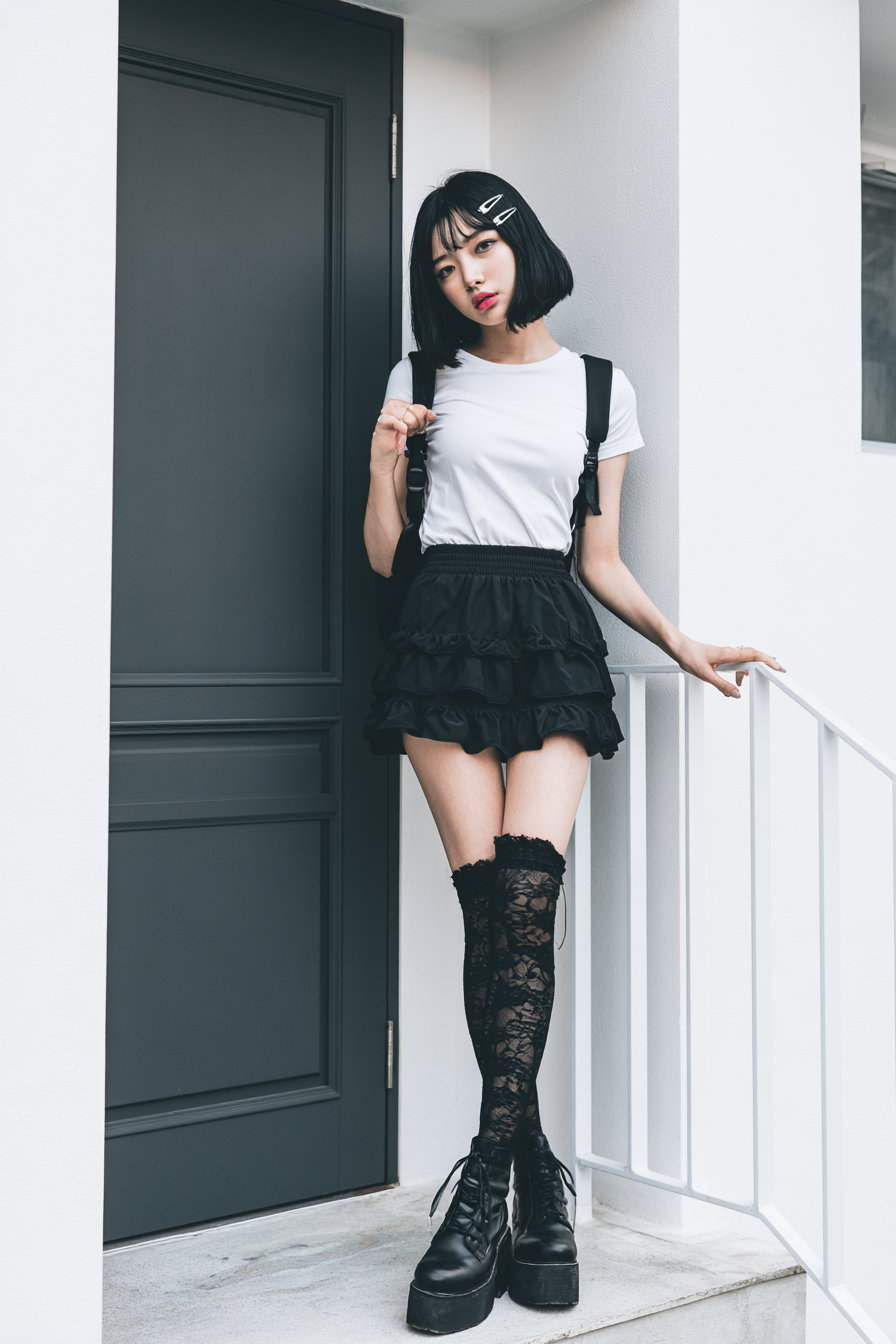 A young woman with short black hair stands on a doorstep wearing a white t-shirt, layered black mini skirt, lace-patterned thigh-high socks, and black platform boots, leaning against a white railing next to a dark gray door