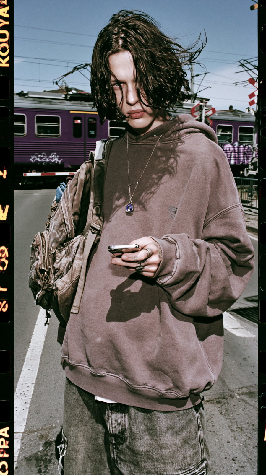 A young person wearing an oversized brown hoodie and multiple rings stands on a city street holding a phone, with a purple train and power lines in the background.
