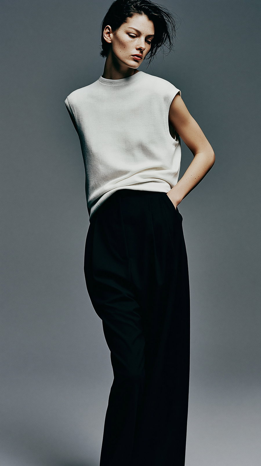 Portrait of a person with short dark hair wearing a sleeveless white top and black pants against a gray background