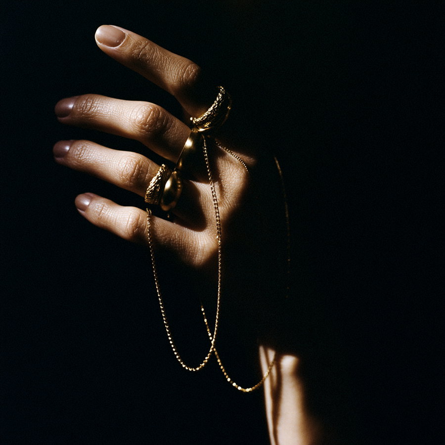 A hand with golden rings on fingers and a fine golden chain, illuminated against a dark background