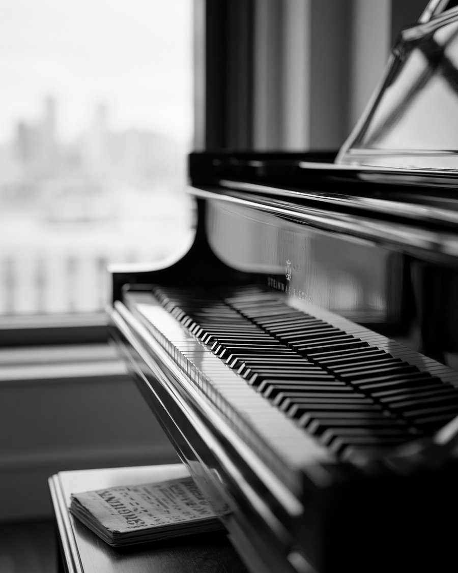 Black and white photo showing a grand piano keyboard with music sheets underneath, window in the background