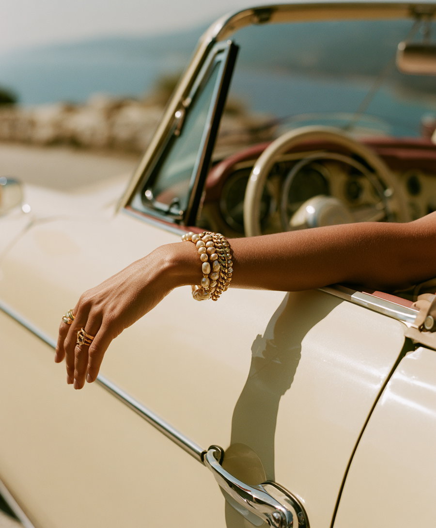 A person's arm with bracelets and rings extended out of a beige vintage convertible car door with blurred background.