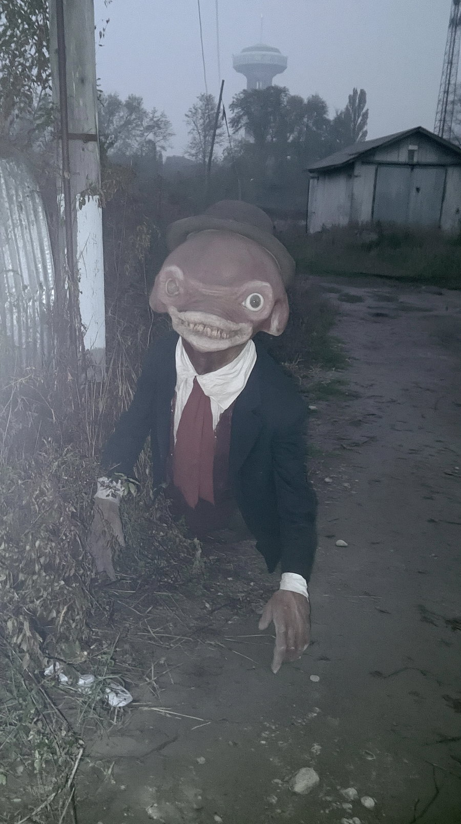 A humanoid figure with a distorted face, large eyes, and sharp teeth wearing a suit, hat, and red tie crawling on a dirt path near dry vegetation and rustic buildings in foggy conditions