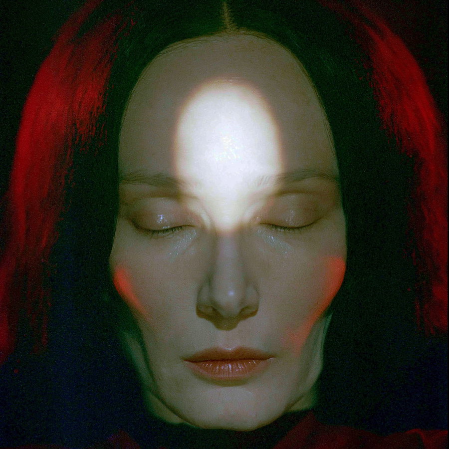 A close-up portrait of a person with eyes closed in soft lighting, face partially illuminated with red and white light accents on dark background
