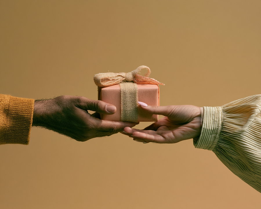 A close-up image of two hands, one handing over a gift wrapped in pink paper and beige ribbon to another, set against a plain beige background
