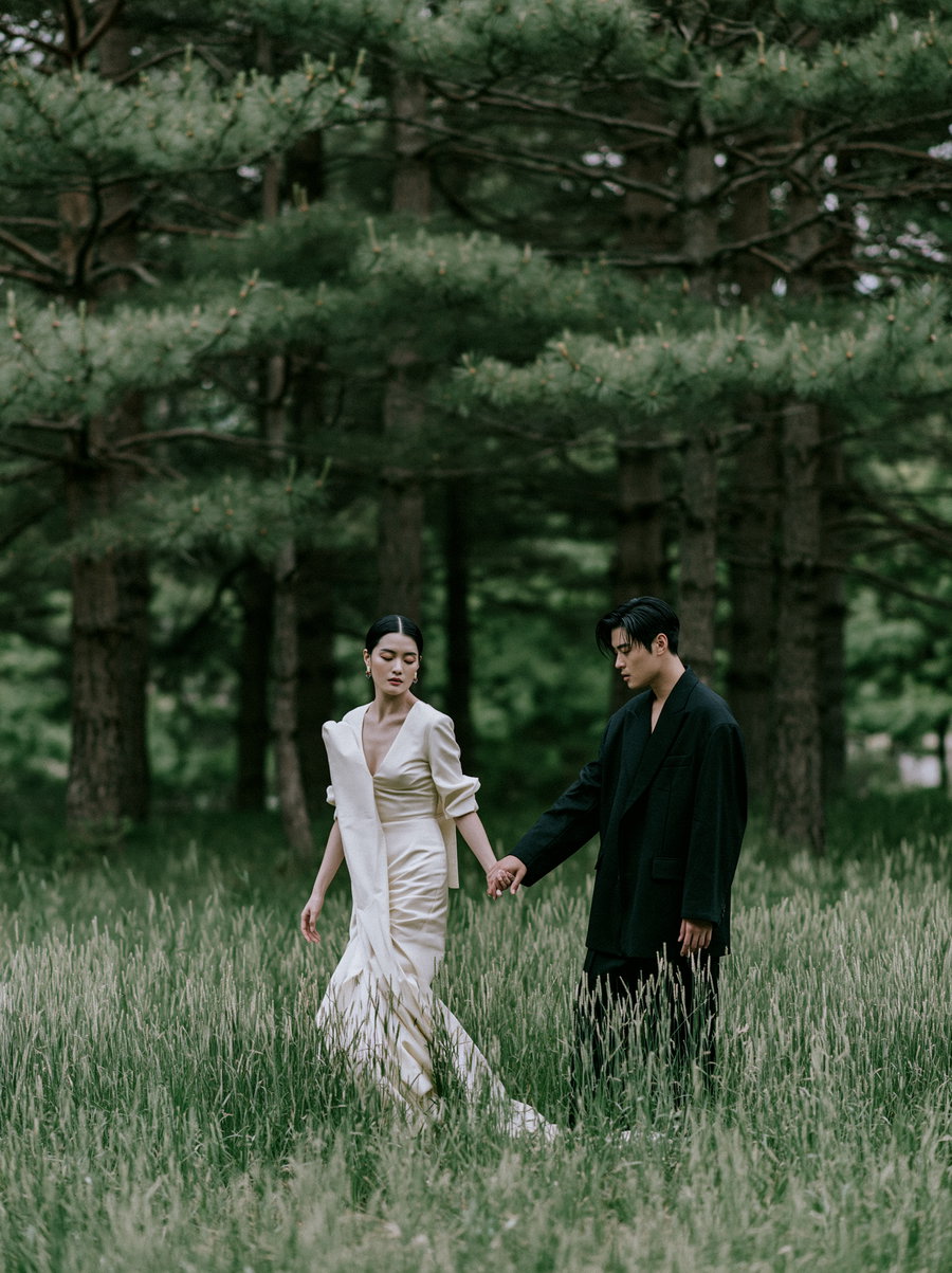 A young man and woman in stylish clothing holding hands and walking through tall grass in a dense forest with pine trees in the background