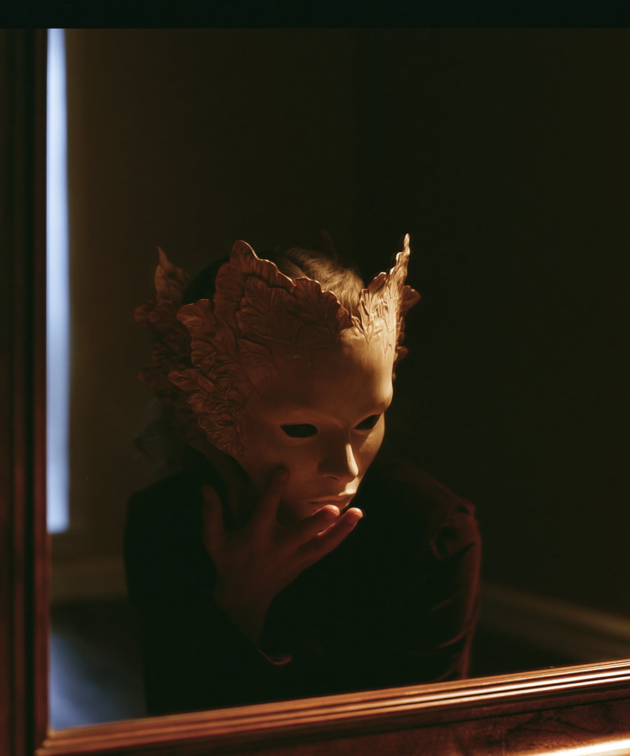 A person with a golden mask with leaf-like structures covers their face, touching the mask with one hand while looking into a mirror in a dark room with soft light illuminating the mask