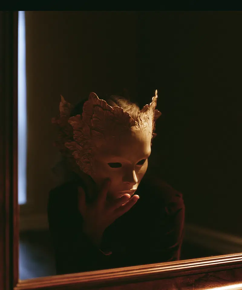 Person wearing a detailed, leaf-patterned mask reflected in a mirror in dim light