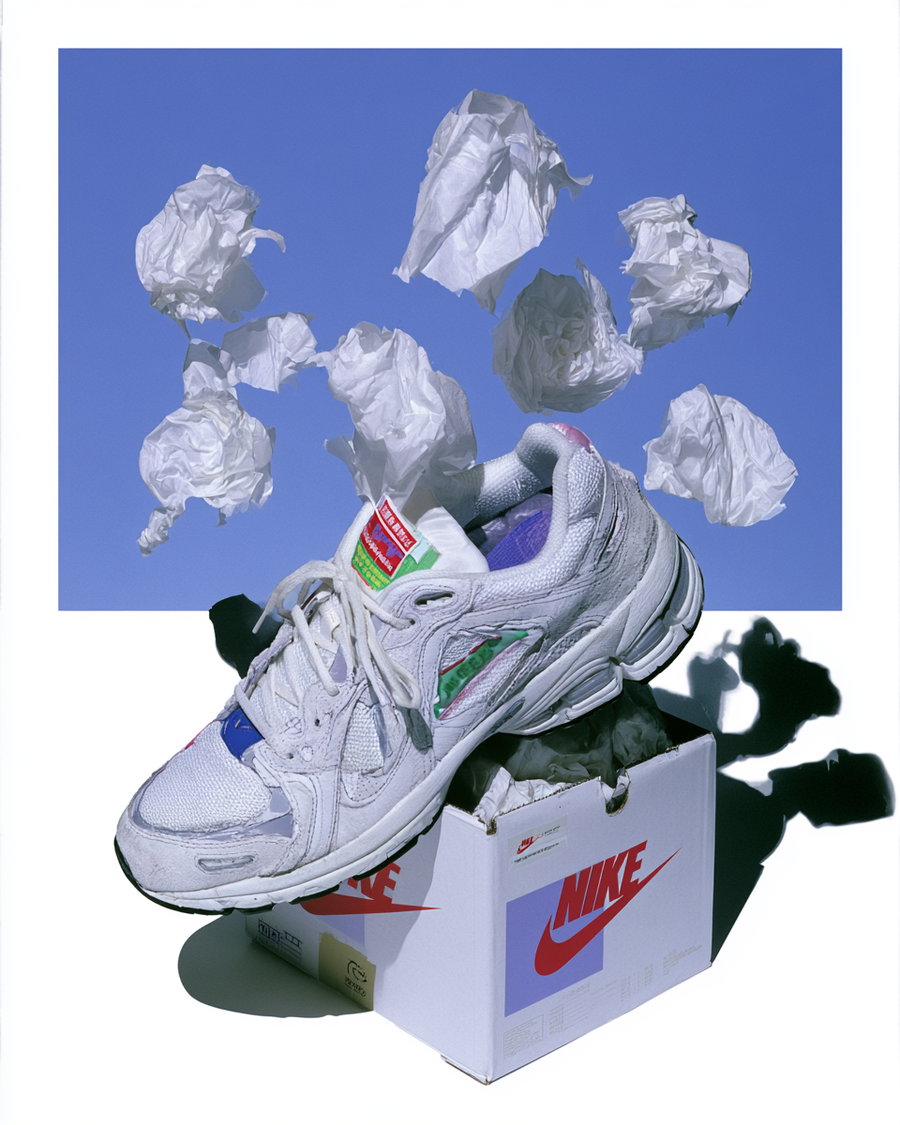 A white Nike sneaker placed on top of a Nike shoe box with several crumpled pieces of white paper floating above against a blue background