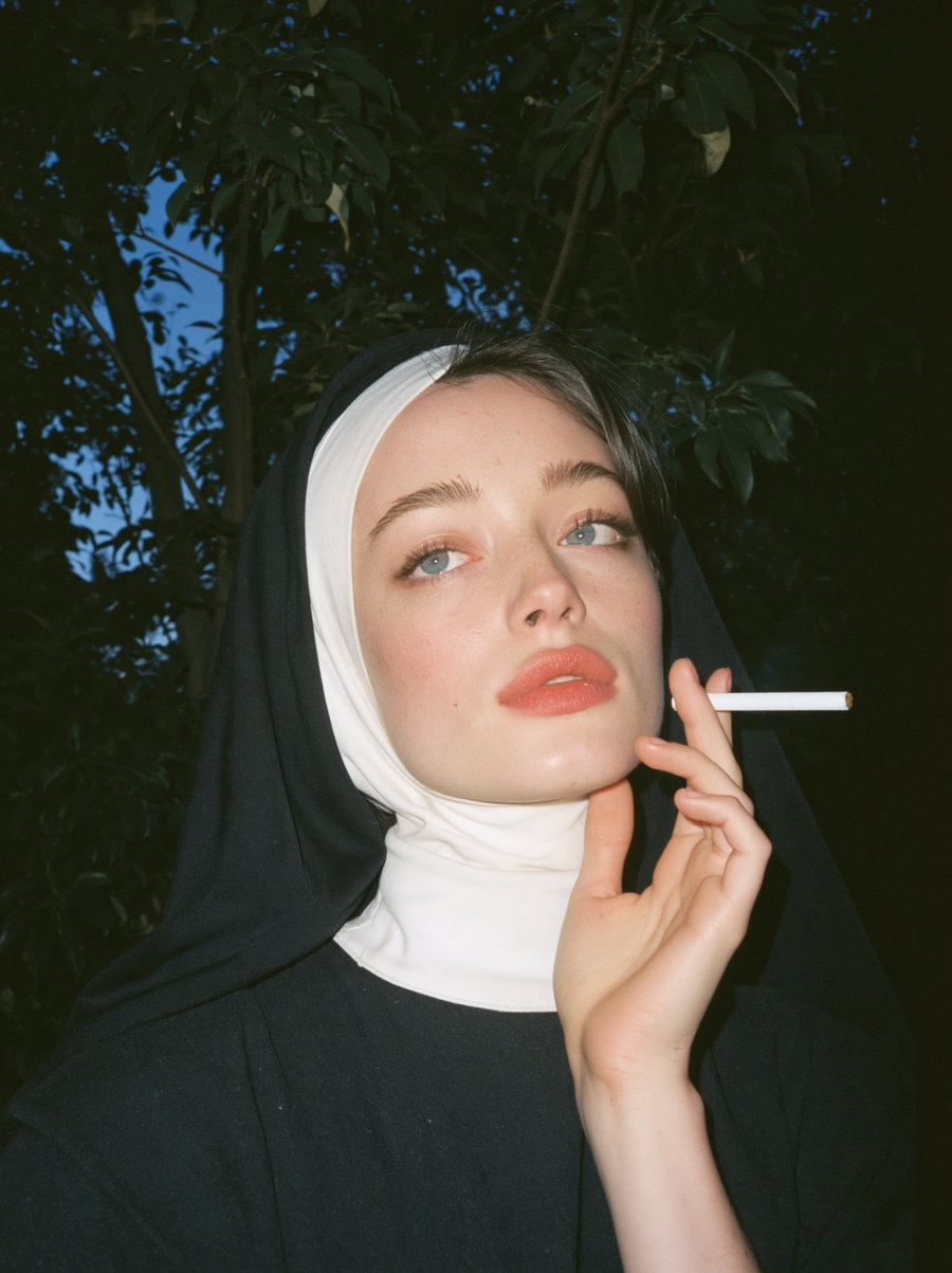 Close-up portrait of a woman wearing a black and white nun's habit, holding a cigarette near her face with dark green leaves in the background
