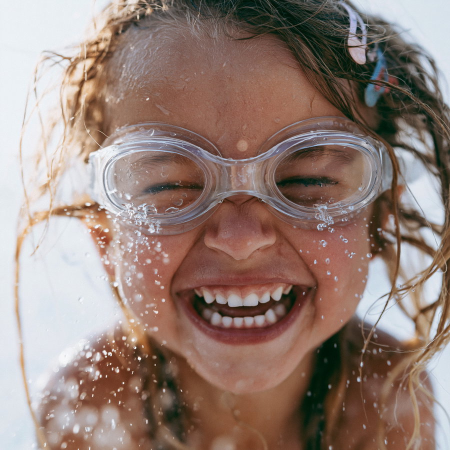 A close-up photo of a child with wet hair wearing clear swimming goggles, laughing with eyes closed and water droplets splashing around their face.