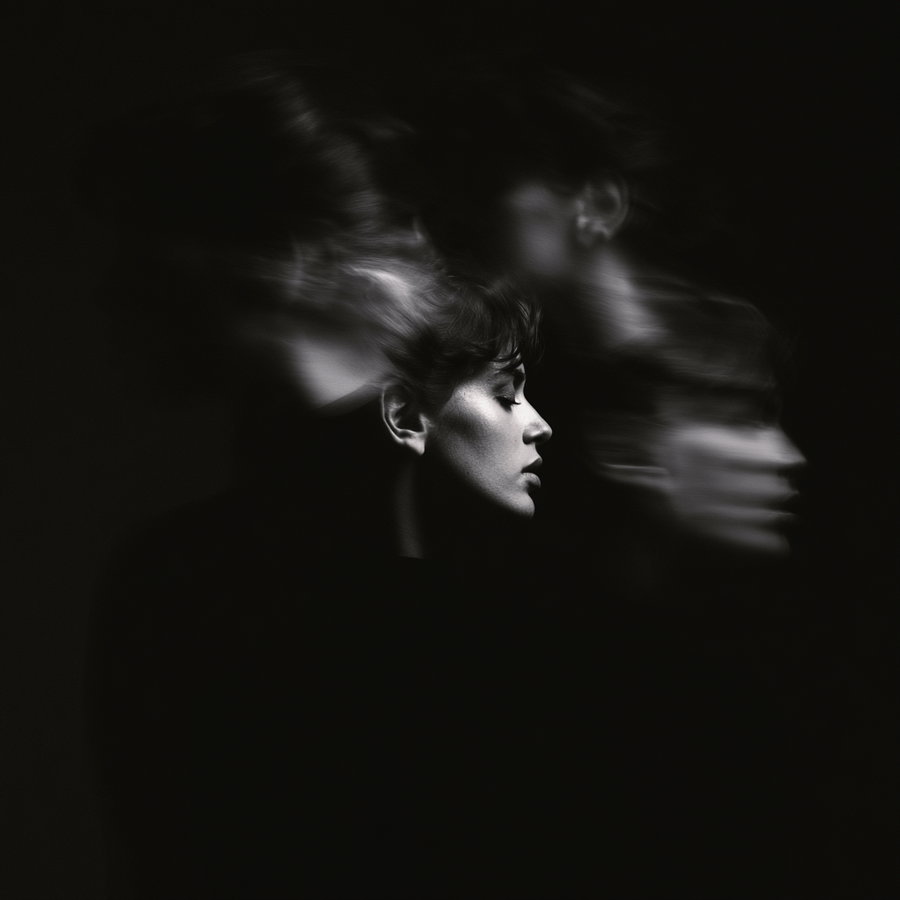 A monochrome artistic portrait of a woman with her profile in focus and multiple motion blurred ghostly exposures around her head on a dark background