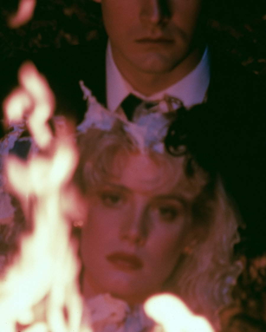 A blurred close-up image of a woman in a white bridal outfit with a man in a suit behind her, both partially obscured by flames in the foreground
