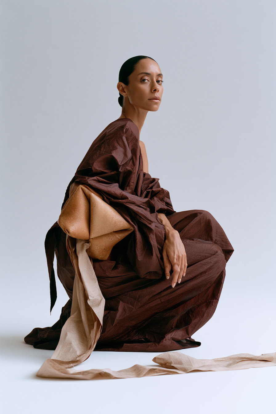 A woman with dark hair tied back is seated against a plain light background, wearing a voluminous brown dress with a textured beige fabric element extending to the floor