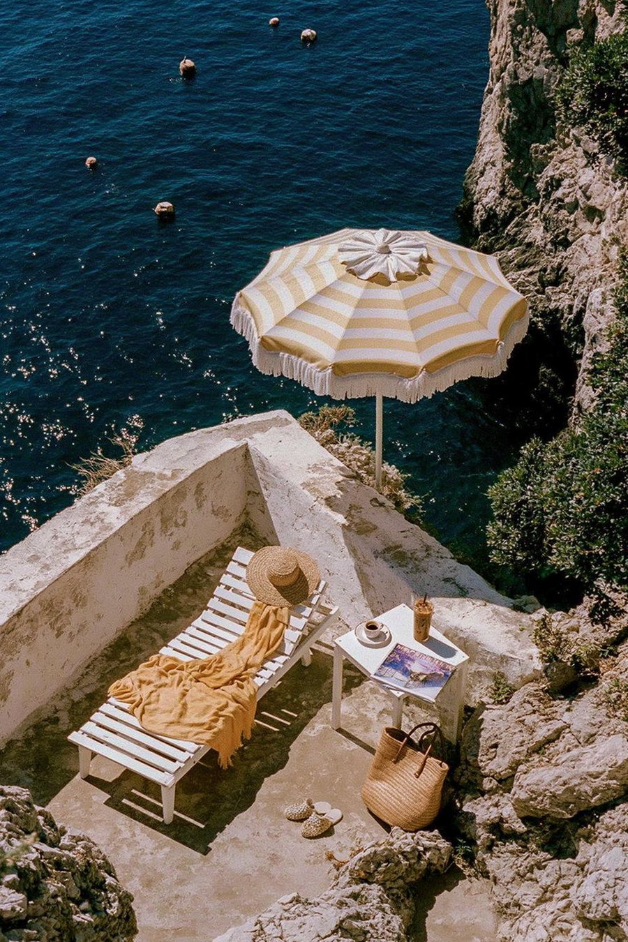 A white lounge chair with a straw hat and yellow blanket on it next to a small white table holding a coffee and iced drink, bordered by rocky cliffs and a striped beach umbrella by the blue ocean
