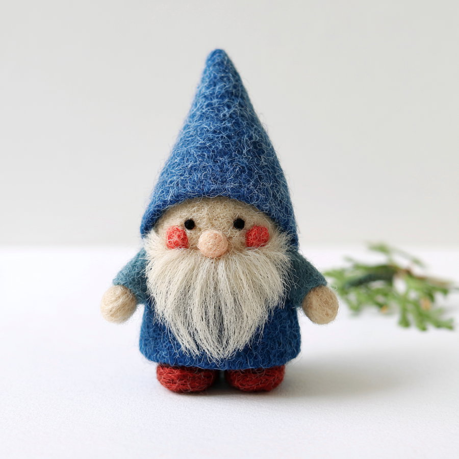 A small handcrafted felt gnome figure with a tall blue hat, white beard, chubby cheeks with red blush, blue clothes, and red shoes standing on a white surface with a blurred green branch in the background