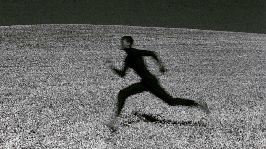 A blurred image of a person in dark clothing running across a grassy field under a dark sky.