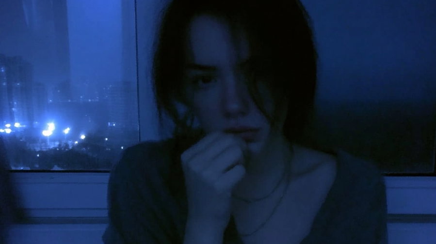 A woman with dark hair partly covering her face sitting indoors by a window at night with blue lighting, looking contemplative with her hand near her lips, city lights visible outside the window.