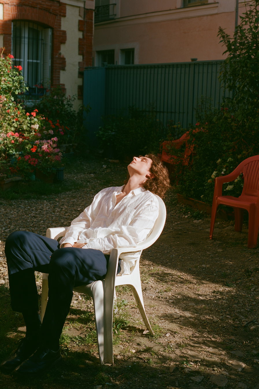 A young man with curly hair wearing a loose white shirt and dark pants sitting on a white plastic chair outdoors, leaning back with eyes closed, surrounded by garden plants and red chairs