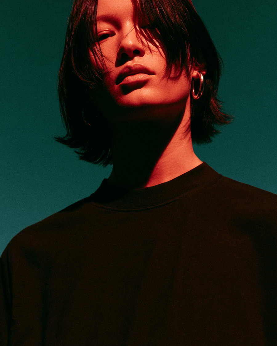Close-up portrait of a man with shoulder-length black hair wearing a black shirt and a large silver hoop earring on a greenish background lit with red light
