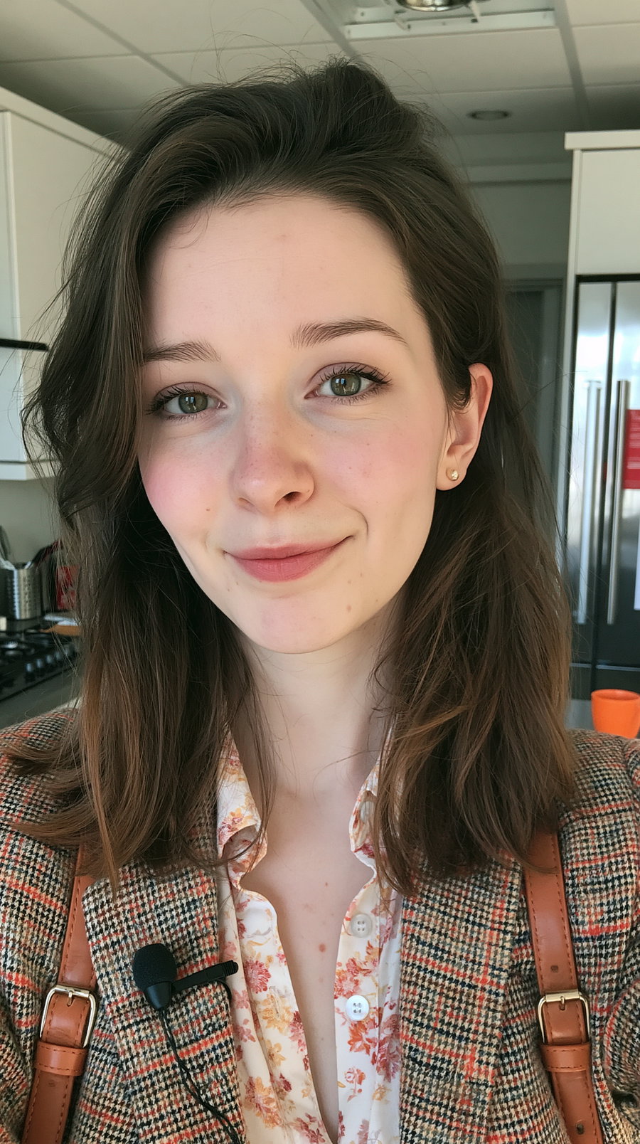 A young woman with shoulder-length brown hair wearing a floral blouse and plaid jacket, smiling softly in a kitchen setting with a microphone clipped to her jacket