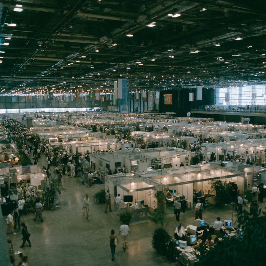 View of a large indoor convention center filled with numerous white booths and partitions, crowded with people walking through aisles, several computer monitors visible in front booths, overhead lights and flags hanging from the ceiling