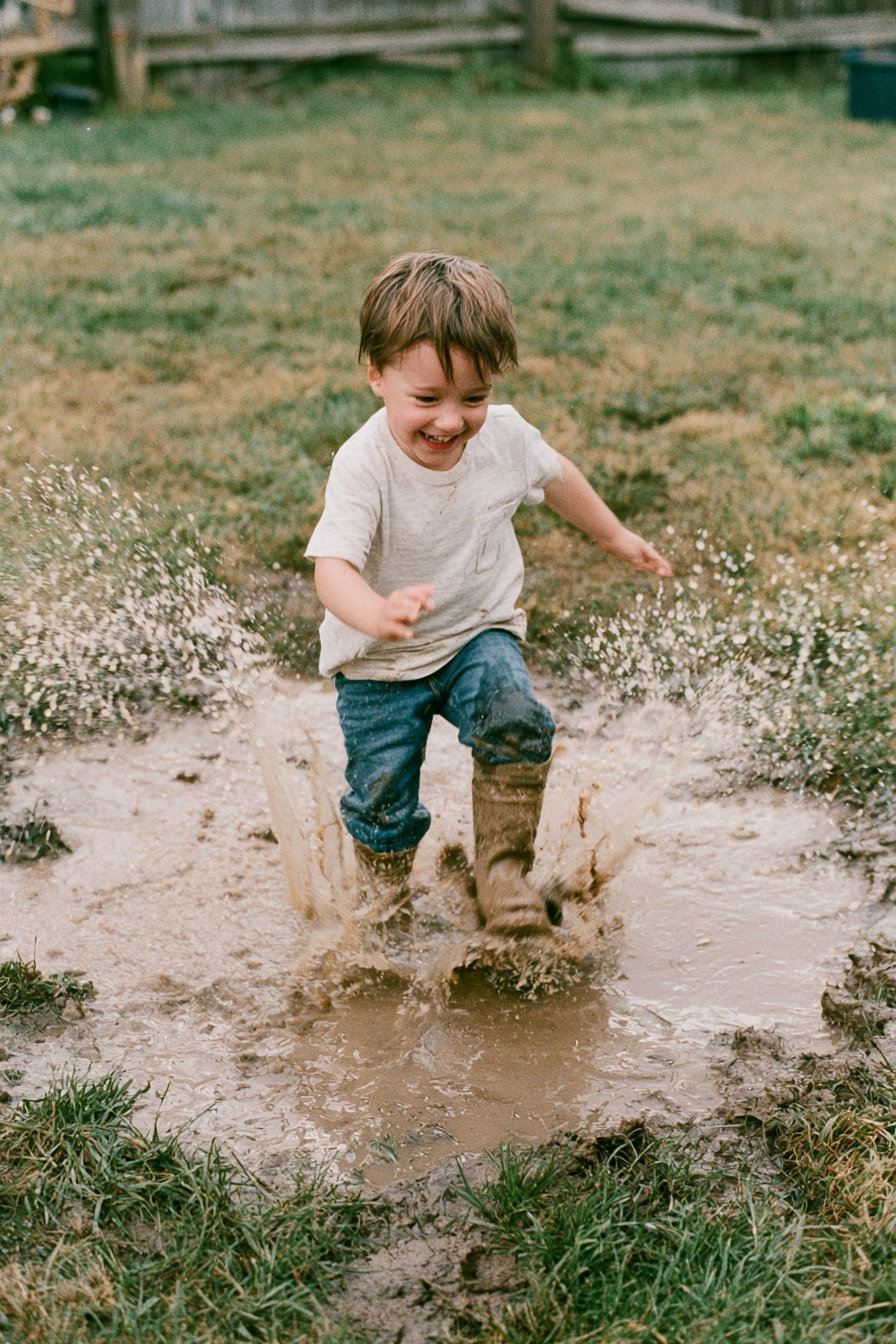 A happy young boy wearing a white t-shirt, jeans, and brown boots is running and splashing through a large muddy puddle on a grassy field.