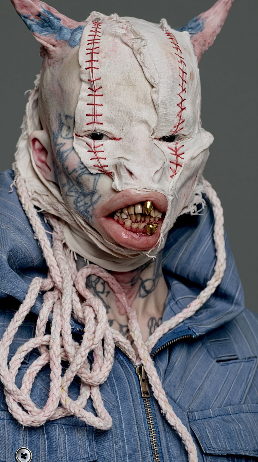 A close-up portrait of a person wearing a grotesque mask made of stitched fabric with horns, revealing golden teeth and tattoos on the face and neck, dressed in a blue jacket with braided rope around the neck.