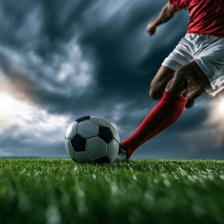 Close-up of a soccer player wearing red socks and white shorts kicking a black and white soccer ball on a green grass field with a cloudy sky background
