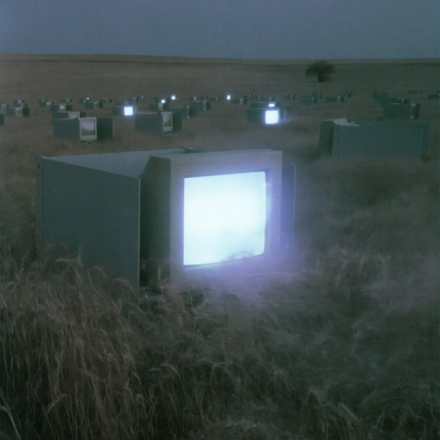 A field of dry grass covered in mist, scattered with many vintage CRT televisions displaying bright white glowing screens, under a darkening sky.