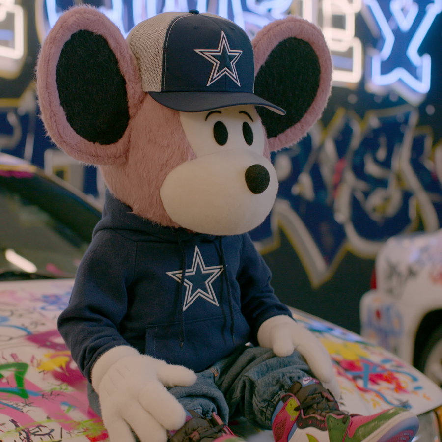 Large mouse character in a blue hoodie and cap with a star logo, sitting on colorful graffiti-covered car in an urban setting