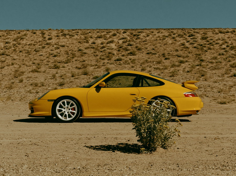 A yellow vintage sports car with white rims on a dry, sandy road with a small bush in the foreground and barren hillside in the background under a blue sky