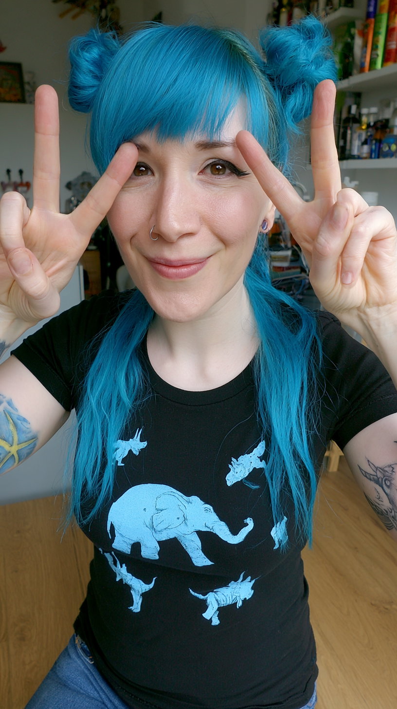 A smiling woman with bright blue hair styled in two buns, wearing a black t-shirt with blue animal prints, showing peace signs with both hands in front of a room background