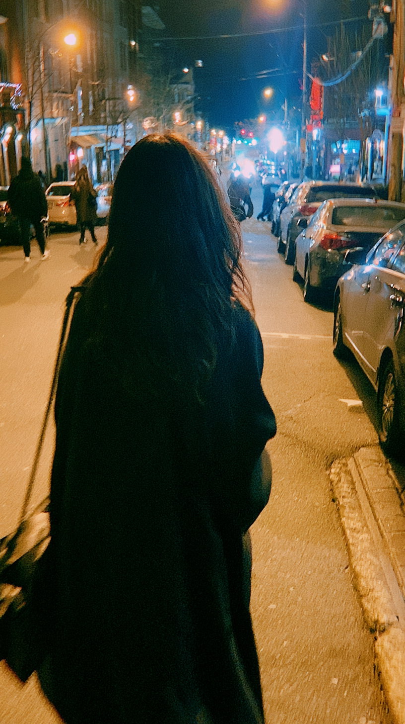 A person with long dark hair and wearing a dark coat walks down a street at night with cars parked on the side and bright street lights illuminating the scene.