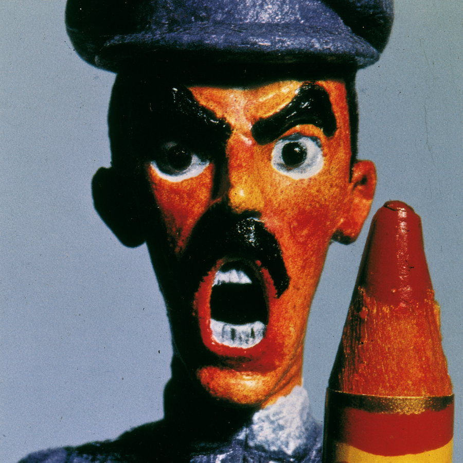 A close-up image of a puppet with an angry expression, painted orange face, black mustache, thick black eyebrows, wide open mouth showing teeth, and wearing a dark cap, holding a large red pencil