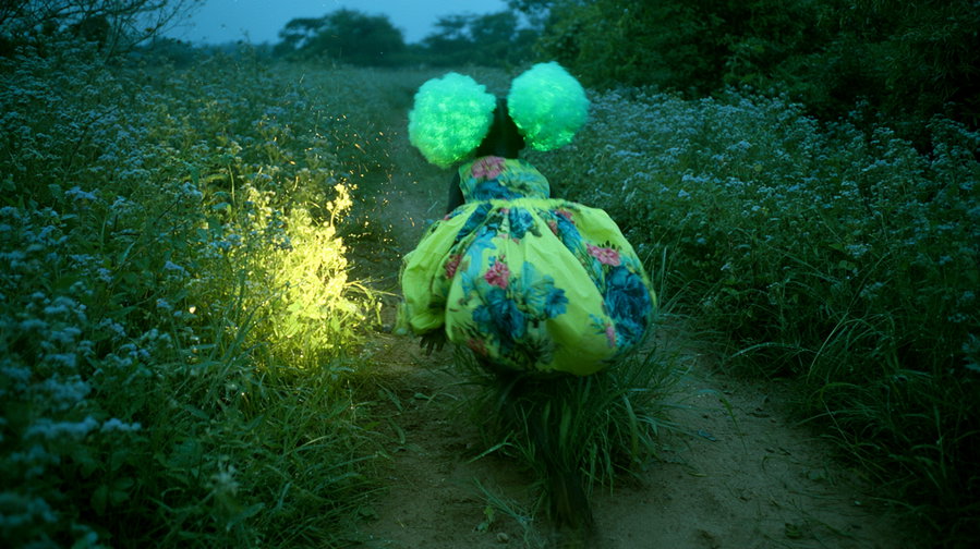 A figure walking on a dirt path through tall green plants with small light blue flowers, wearing a bright yellow floral puff dress and large glowing green headpieces, with a soft glowing light illuminating the foliage on the left side.