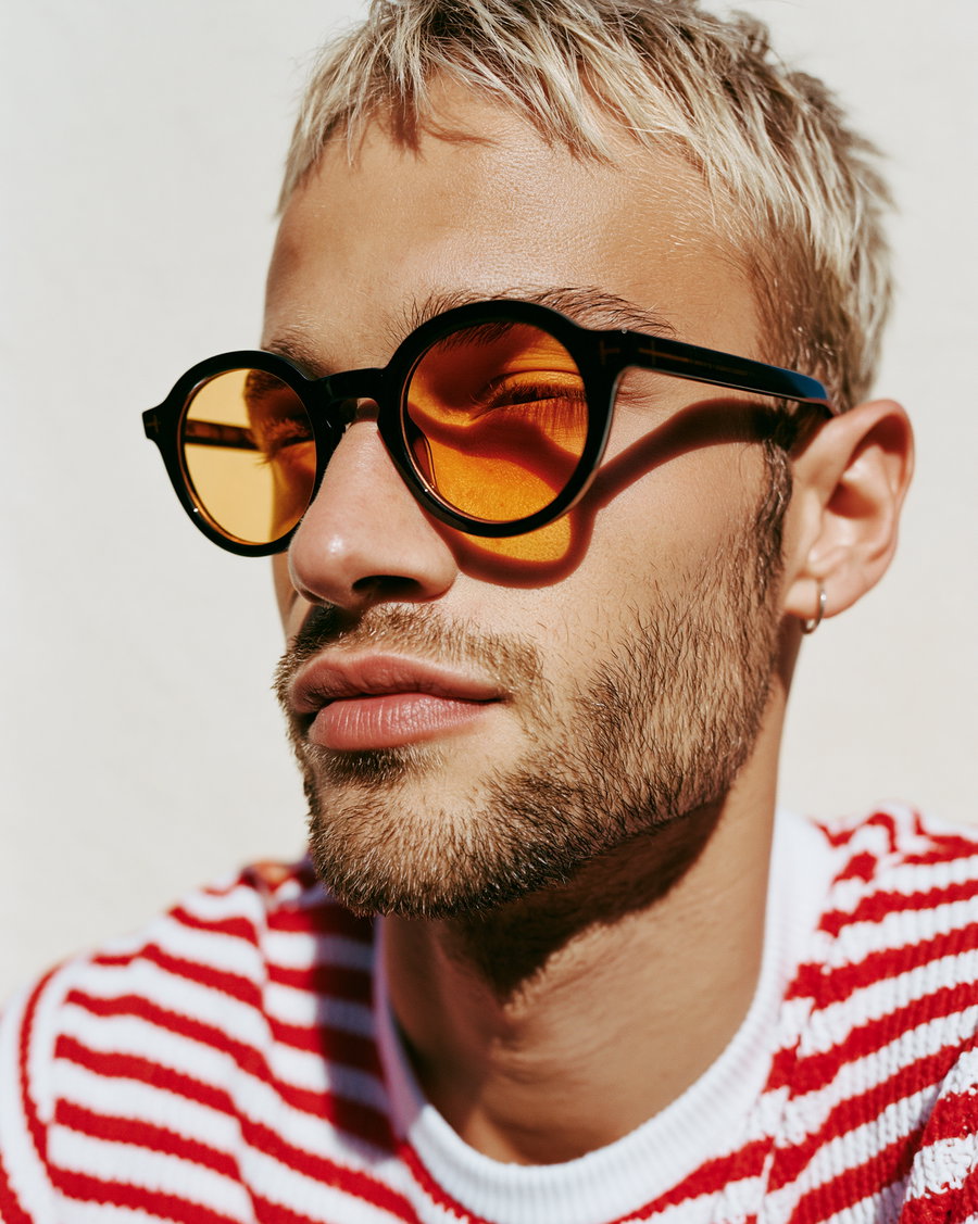 A man with short blond hair and beard wearing black-framed round orange sunglasses, a small hoop earring, and a red and white striped sweater against a plain light background