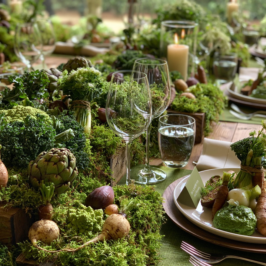 A close-up of a dining table decorated with fresh vegetables, moss, glassware, candles, and plates with green sauces, arranged on a wooden table with green cloth placemats
