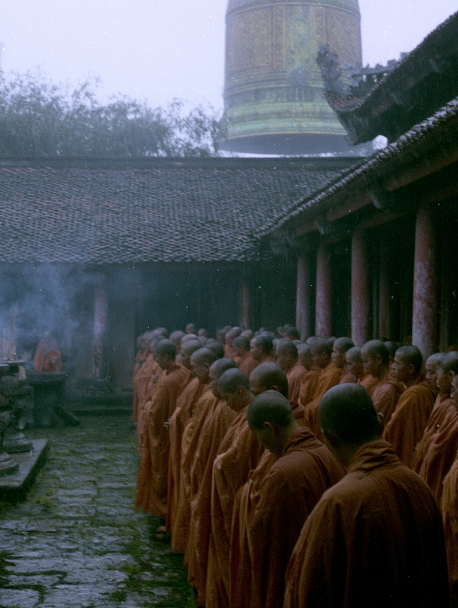 A large group of monks in orange robes standing quietly in a stone courtyard with moss-covered ground, near traditional buildings and a large ornate bell in the background