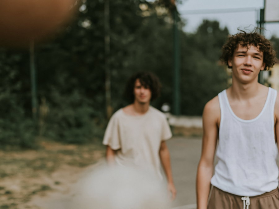 Two young men with curly hair, one in a white tank top and brown shorts and the other in a light-colored t-shirt, standing on an outdoor sports court with trees and greenery behind them