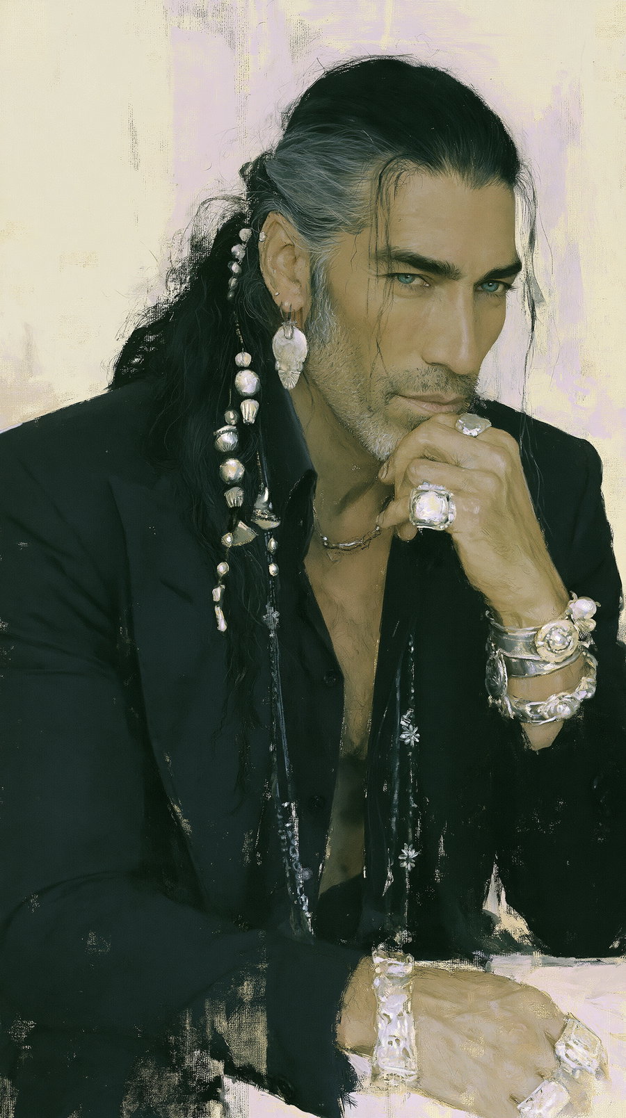 A man with long dark hair tied back, wearing multiple silver rings, bracelets, and an earring. He is dressed in a black jacket and shirt, resting his hand on his chin with an intense gaze.
