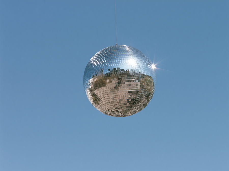 A shiny disco ball made of small mirrored tiles hanging from a thin wire with a bright sun reflection and clear blue sky behind it