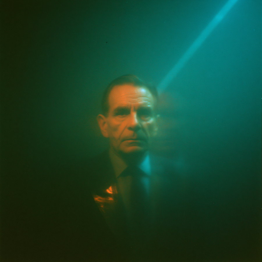 A moody portrait of a serious middle-aged man in a suit, surrounded by green and teal lighting that creates a mysterious and dark atmosphere.