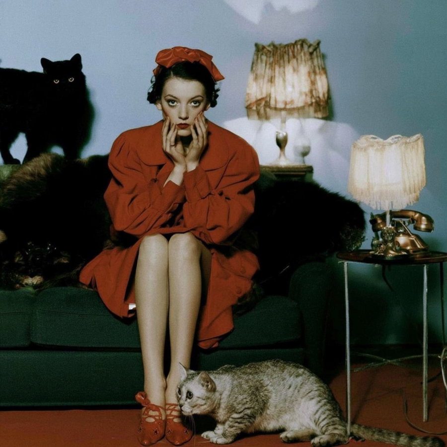A woman wearing a red dress and headband is sitting on a green couch, surrounded by a black cat on the couch and a gray tabby cat on the floor. Two vintage lamps and a rotary phone on a side table are also visible.