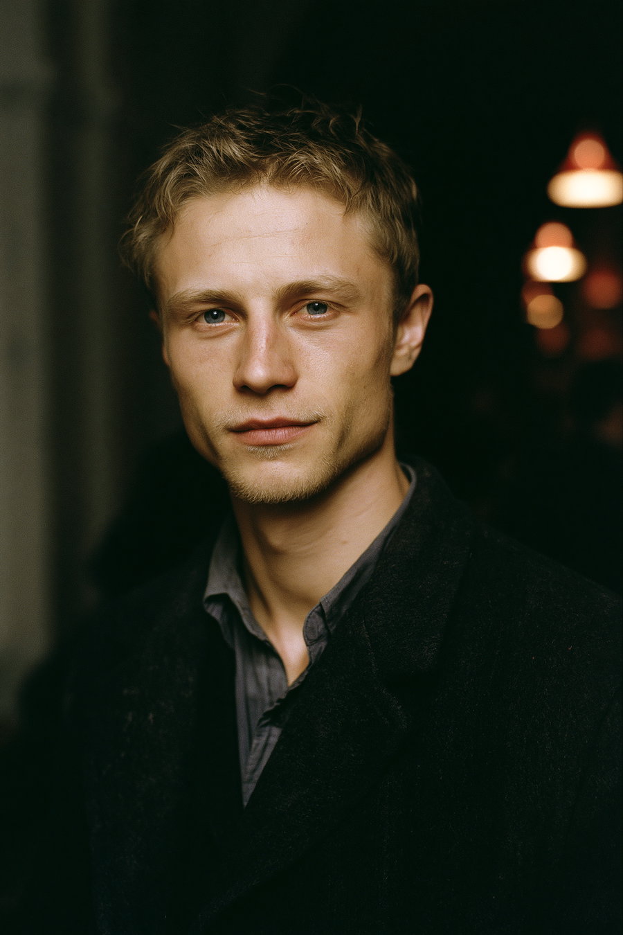 Close-up image of a young man with short blond hair, wearing a dark coat and a gray shirt, set against a dark background with some out-of-focus lights