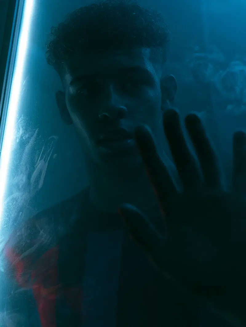 Moody portrait of a young man with curly hair touching a glass pane illuminated by blue light