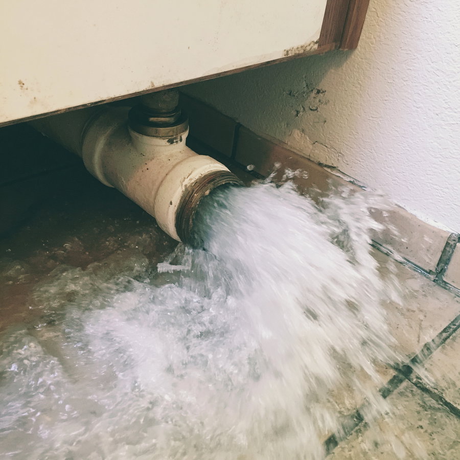 A large volume of water forcefully flowing out from a pipe beneath a cabinet onto a tiled floor with some wall damage visible nearby.