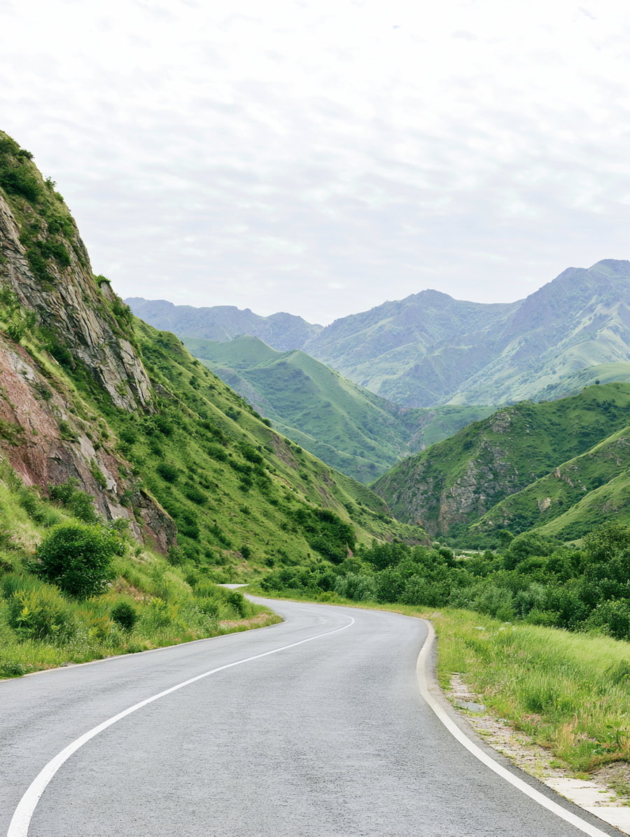 A paved road winding through a valley surrounded by green mountain slopes and vegetation, with a cloudy sky overhead