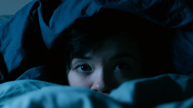 Person partially hidden under blue covers with eyes wide open in dim light